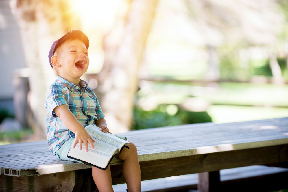 Little boy with purple cap and blue plaid shirt laughing while sitting on a bench with a book open on his lap